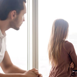 A man, forlorn, looking at a woman who is sitting with her back to him and her hands crossed.