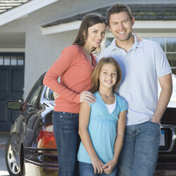 A family of three standing outside of their home with their car in the background.