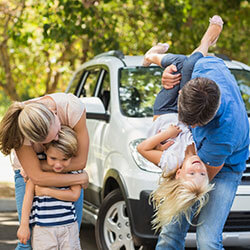 A family hugging and being playful with the family car in the background.