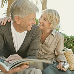 An older man and woman taking a break while travelling. He's looking at a travel brochure while she holds her camera.