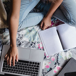 An overhead photo of a teenager sitting on her bed studying and researching online with her laptop.